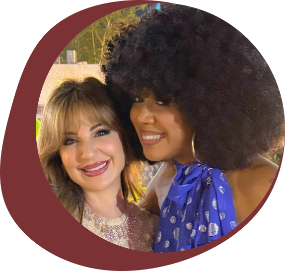 Two women smiling together, one with curly hair and a blue dress, the other with straight hair and a light outfit.